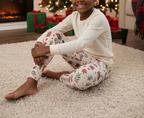 A child sitting on a cream shag rug in a cozy living room, wearing a cream long-sleeve top and matching cream leggings with a festive red, green, and gold holiday print. A Christmas tree and wrapped gifts are visible in the background.