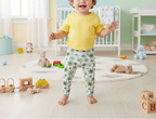 Child in a yellow shirt and floral pants standing in a nursery room with toys and furniture.
