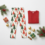 A flat-lay holiday outfit set featuring a pair of cream leggings with a red and green Christmas tree pattern, and a folded red long-sleeve shirt with white speckles, styled with pine branches, ornaments, and gift boxes on a white background.