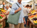 Man holding a striped reusable bag in a bustling market with fresh produce.