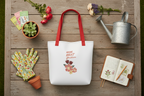 A white tote bag with red handles and a floral graphic reading 'MOM BEST Gardeners' is arranged on a rustic wooden surface with gardening props including gloves, a watering can, flowers, a notebook, and small pots.