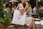 Woman sitting outdoors with a tote bag and drink, surrounded by people and greenery.