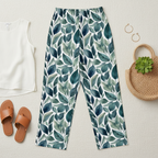 Leaf-patterned pants on a light surface with sandals, a bag, and a plant.