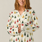 A woman wearing a long-sleeve button-up pajama shirt with a festive Christmas tree pattern in red, green, and mustard yellow on an off-white background, set against a neutral textured backdrop.