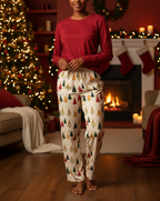 A woman stands barefoot in a cozy living room wearing a festive Christmas pajama set. The set includes a solid red long-sleeve top and cream-colored pants decorated with a repeating pattern of small Christmas trees in green, red, and gold.
