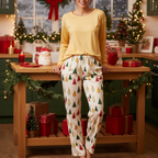 A woman wearing a yellow long-sleeve top and cream holiday pajama pants with a Christmas tree print, standing in a decorated Christmas kitchen behind a wooden table with wrapped gifts.