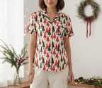 A woman wearing a short-sleeve button-up pajama shirt with a Christmas tree pattern in red and green on an off-white background, paired with cream pants, standing in a decorated holiday room.