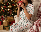 A woman in a white Christmas-themed pajama set with a tree print sits on a tufted chair holding a red mug, with a decorated Christmas tree and wrapped gifts in the background.