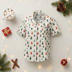 A short-sleeve pajama top with a Christmas tree print, laid flat on a light beige fabric background decorated with holiday props including a mug of marshmallows, a small gift box, pine branches, cinnamon sticks, and fairy lights.