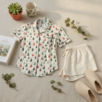 A children's pajama set laid flat on a light fabric surface, including a short-sleeve button-front pajama shirt with an allover Christmas tree print and matching cream shorts. A mug, small potted succulent, book, and slippers are arranged around it as styling props.