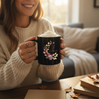 Person holding a mug with floral design and whipped cream, sitting in a cozy room.