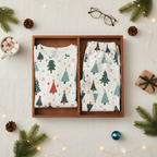 A flat-lay of a matching Christmas-themed pajama set in a wooden box, featuring a white long-sleeve top and pants with a colorful tree pattern, surrounded by holiday props like pine branches, pinecones, glasses, and a mug of hot chocolate.