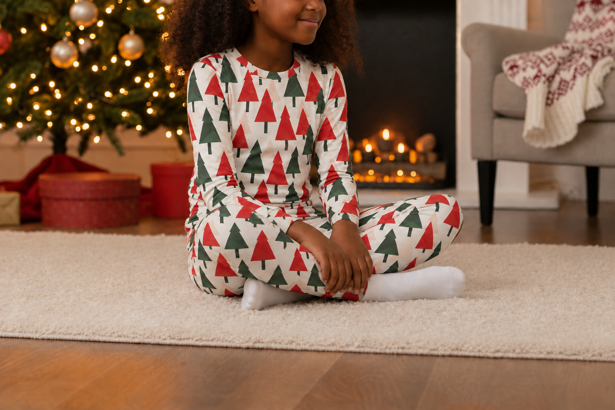 A child sitting cross-legged on a white rug in a cozy living room, wearing a long-sleeve pajama set with a red and green Christmas tree pattern on a white background. A decorated Christmas tree, fireplace, and armchair are visible in the background.