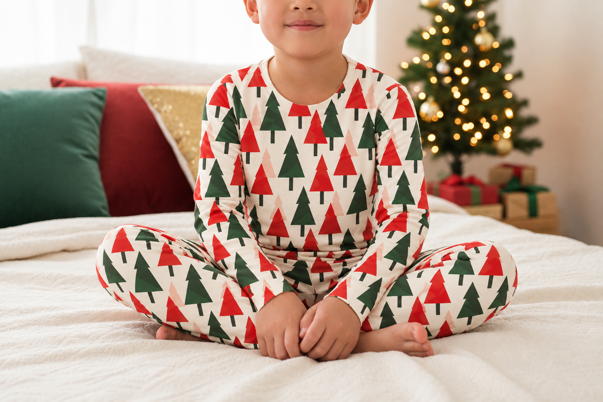 A child sitting cross-legged on a bed wearing a long-sleeve pajama set with a repeating red and green Christmas tree pattern on a white background. A decorated Christmas tree and wrapped gifts are visible in the softly lit bedroom background.