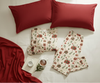 Christmas Present men’s pajama top and pocket-free pants arranged on white bedding with a red throw and cocoa