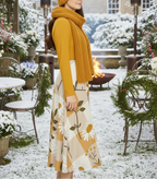 Woman in mustard yellow sweater and floral skirt standing in a snowy outdoor setting with a fire pit and Christmas decorations.