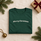 Folded green Merry Christmas tee with greenery, a brass bell and kraft gift