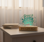 Decorative sign with 'Grow a Little Cheer' text on a table with light curtains in the background