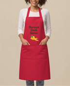 A woman wearing a bright red apron over a white long-sleeve shirt and gray jeans, standing against a plain beige background. The apron has the words 'Harvest Cook Savor' in black script with a small yellow and red pepper graphic below.