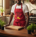 A smiling man in a kitchen wearing a red apron with the text 'Harvest Cook Savor' and a yellow pepper graphic. He is standing behind a wooden cutting board on a wooden counter with potted herbs in a warm, well-lit kitchen.