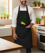 A man wearing a black kitchen apron with green text reading 'Harvest Cook Savor' and a yellow lemon graphic, standing in a bright modern kitchen.