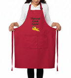 A person wearing a red apron with the words 'Harvest Cook Savor' and a small yellow pepper graphic on the chest, holding the sides of the neck strap against a plain white background.
