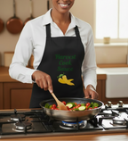 A woman wearing a black kitchen apron with green and yellow 'Harvest Cook Sauce' text and a pepper graphic, stirring a pan of vegetables on a gas stove in a kitchen.
