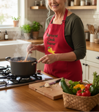 A smiling woman in a kitchen wearing a red apron with the words 'Harvest Cook Savor' and a yellow bird graphic. She is stirring a pot on a gas stove, with fresh vegetables on a cutting board and basket in the foreground.