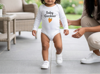 A toddler wearing a white long-sleeve bodysuit with the text 'Baby Gardener' and a small orange leaf graphic walks outdoors while being supported by an adult. The background shows a patio with a wicker chair and a blurred garden.