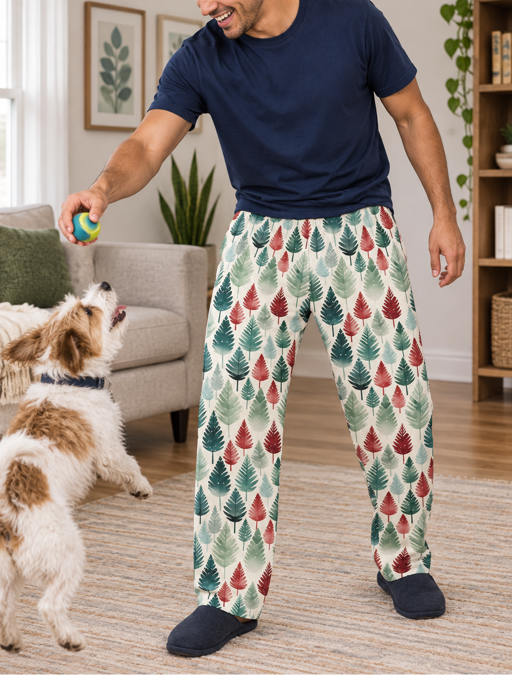A man wearing a navy blue t-shirt and patterned pajama pants with a green and red leaf design, playing with a small dog in a bright living room.