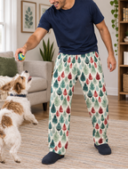 A man wearing a navy blue t-shirt and patterned pajama pants with a green and red leaf design, playing with a small dog in a bright living room.