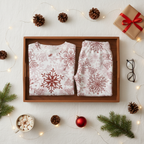 Folded Red Snowflake men’s pajama top and matching pocketless pants in a wooden split tray with cocoa, greenery, gifts, and warm lights