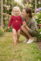 A toddler in a red long-sleeve bodysuit with 'Baby Grandpa' and a small yellow sun graphic walks barefoot in a grassy garden path while a woman crouches beside her, surrounded by blooming pink roses.
