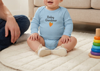 A baby with red hair wearing a light blue long-sleeve bodysuit with 'Baby Gardener' and a carrot graphic, sitting on a cream quilted blanket in a living room.