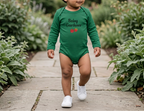 A toddler walking on a stone garden path wearing a green long-sleeve bodysuit with 'Baby Gardener' and tomato graphics, paired with white sneakers.