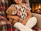 Child in Christmas Tree kids pajama top in an armchair with a teddy bear by the fireplace