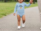 A toddler walking outdoors on a paved path while holding an adult's hand. The child is wearing a light blue long-sleeve bodysuit with the words 'Baby Gardener' and a small orange heart graphic on the front.