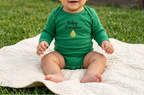 A baby sitting outdoors on a white blanket wearing a green long-sleeve bodysuit with 'Baby Gardener' and a pear graphic printed on the chest.