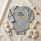 A heather gray long-sleeve baby bodysuit with 'Baby Gardener' and a carrot graphic printed on the chest, styled in a flat lay on a cream fabric background with a plush bunny, cream knit booties, and wooden baby toys.
