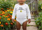 A toddler stands in a garden wearing a white long-sleeve bodysuit with the text 'Baby Gardener' and a small pear graphic on the chest. The background shows colorful flowers, green plants, and a wooden fence.