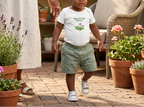 Toddler wearing white New to the Patch flower onesie on a patio.