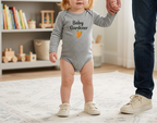 A toddler girl wearing a gray long-sleeve bodysuit with 'Baby Gardener' and a carrot graphic stands on a rug holding an adult's hand in a bright nursery setting.