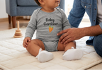 A baby wearing a heather gray long-sleeve bodysuit with 'Baby Gardener' and a carrot graphic, sitting on a cream quilted mat with a man in a denim shirt next to them in a bright nursery.