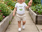 Toddler wearing white New to the Patch flower onesie in a garden.