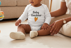 A baby wearing a white long-sleeve bodysuit with 'Baby Gardener' and a carrot graphic sits on a play mat in a living room while an adult sits beside them.