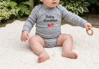 A baby with red hair sits on a white textured blanket in a garden setting, wearing a light gray long-sleeve bodysuit with 'Baby Gardener' and two strawberries printed on the chest, and holding a wooden teething ring.