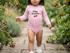 A toddler girl walking on a stone path surrounded by green plants and red berries, wearing a light pink long-sleeve bodysuit with 'Baby Gardener' and a strawberry graphic printed on the front.