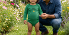 A man in a blue denim shirt and jeans crouches in a garden next to a young toddler wearing a green long-sleeve bodysuit with 'Baby Gardener' printed on the chest. The toddler is smiling and holding the man's hand, with gardening gloves and a small trowel on the grass nearby.