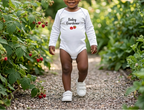 A smiling toddler walking on a gravel path in a garden, wearing a white long-sleeve bodysuit with 'Baby Gardener' and strawberry graphics on the front.