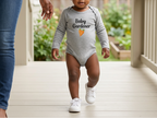 A toddler walking on a porch while wearing a light gray long-sleeve bodysuit with the text 'Baby Gardener' and a small orange leaf graphic. An adult's hand and leg are visible holding the child's arm.